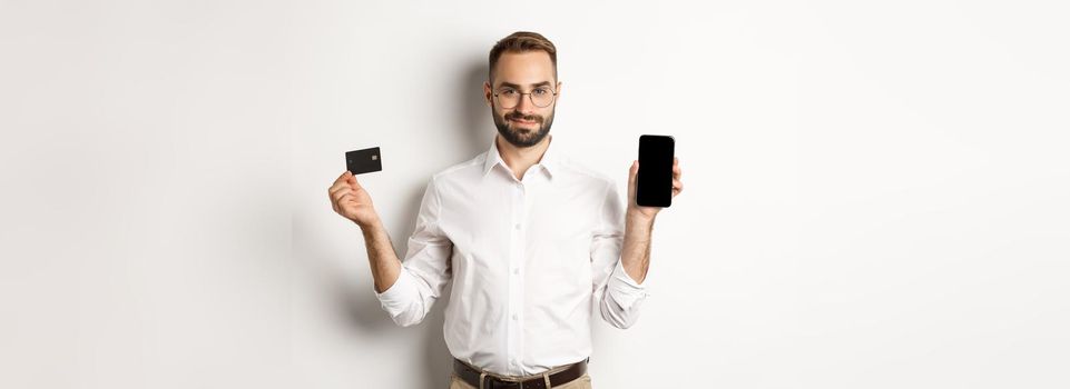 Handsome bearded man showing mobile phone and credit card, shopping online, standing over white background.