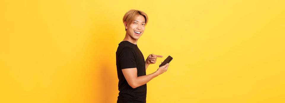 Portrait of pleased smiling handsome asian man, standing in profile and pointing finger at smartphone, recommend app, standing yellow background.