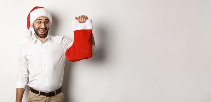 Merry christmas, holidays concept. Happy adult man receive gifts in xmas sock, looking excited, wearing santa hat, white background.