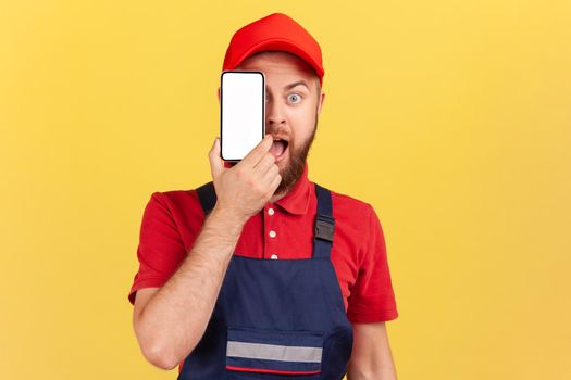 Portrait of shocked amazed bearded worker man in blue uniform standing and covering eye with cell phone with empty display for promotional text. Indoor studio shot isolated on yellow background.