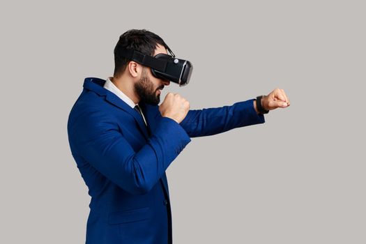 Side view of man virtual reality glasses on head playing fighting game, holding clenched fists up ready to boxing, wearing official style suit. Indoor studio shot isolated on gray background.