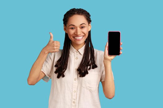 Joyful woman with black dreadlocks smiles happily, points at blank screen of cellualr, advertises new gadget, showing thumb up, wearing white shirt. Indoor studio shot isolated on blue background.