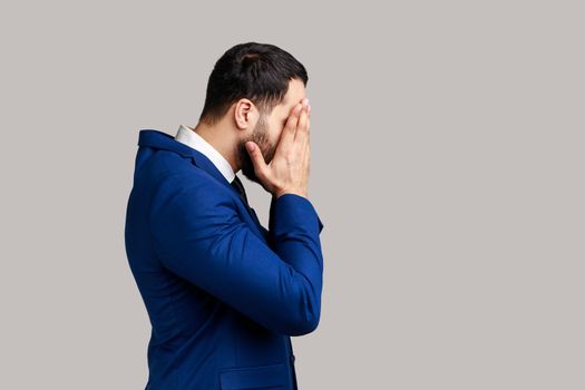 Side view of bearded man closing eyes with hand, dont want to see that, ignoring problems, hiding from stressful situations, wearing official style suit. Indoor studio shot isolated on gray background