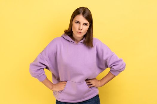 Portrait of young angry serious woman looking at camera with strict negative expression, aggression, keeping hands on hips, wearing purple hoodie. Indoor studio shot isolated on yellow background.