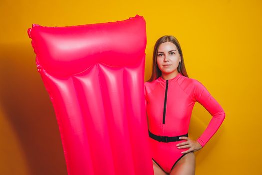 Young slender woman in a pink swimsuit on a yellow background. Girl with an inflatable mattress for swimming in the pool. Summer vacation.