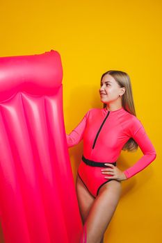 Young slender woman in a pink swimsuit on a yellow background. Girl with an inflatable mattress for swimming in the pool. Summer vacation.