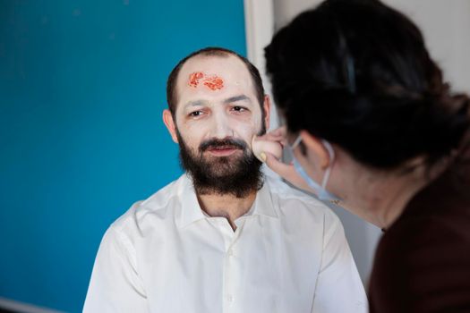 Artist drawing zombie look on man, applying bloody dramatic makeup with scary horror undead costume. Using special effects to create terrible spooky evil corpse face with crazy eyes.