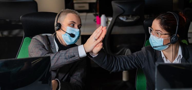 Two business women in masks are giving a high five while sitting at one desk in the office.