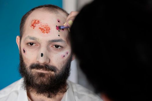 Artist using makeup and costume to create zombie look, sitting in studio. Man waiting to look dramatic, bloody and scary with creepy undead face and frightening eyes. Spooky corpse design.