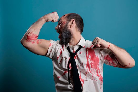 Spooky halloween monster flexing arm muscles on camera, showing biceps and triceps while acting aggressive and dangerous. Crazy wounded zombie eating brain and having bloody scars, fit devil.
