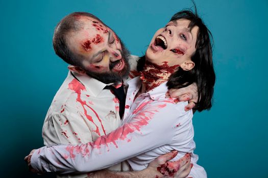 Brain eating halloween zombies hugging over blue background, acting aggressive and frightening. Couple of cruel monsters with blood and dirty scratches sharing hug on camera, bloody wounds.