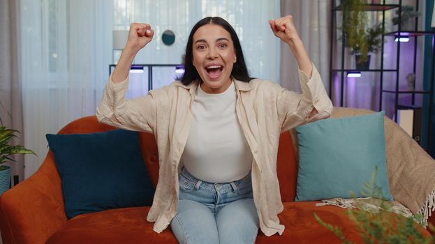 Happy caucasian girl shouting, celebrating success, winning, goal achievement, good news, raising fists in gesture I did it, lottery luck at home apartment. Young woman indoors in living room on couch