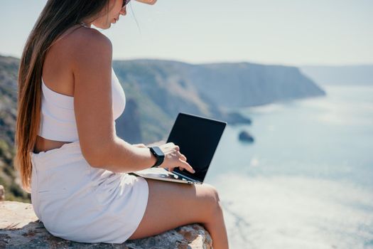 Successful business woman in yellow hat working on laptop by the sea. Pretty lady typing on computer at summer day outdoors. Freelance, travel and holidays concept.