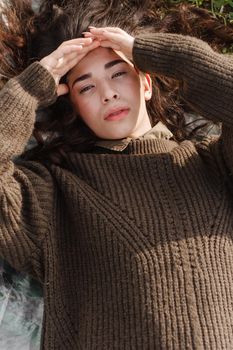 An Asian woman is lying on the ground in a field in a brown sweater. The concept of manufacturing clothing from recycled plastic. An Asian woman is lying on a plastic bag. Portrait of a dark-haired woman with loose hair.