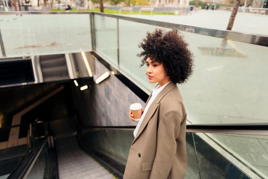 latin business woman entering the subway station on the escalator with a coffee in her hand, concept of urban lifestyle and public transport, copy space for text