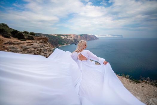 Blonde with long hair on a sunny seashore in a white flowing dress, rear view, silk fabric waving in the wind. Against the backdrop of the blue sky and mountains on the seashore