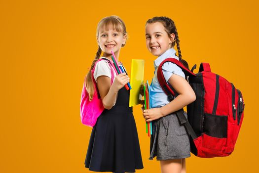 Education Concept. Set portrait of diverse schoolchildren holding stack of academic books and wearing backpack, yellow orange color studio background.