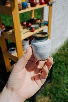 A miniature ceramic jug in a woman's hand. Wooden shelving with ceramic products with your own hands. Green lawn. Market hand made in an open space. Vertical photo.