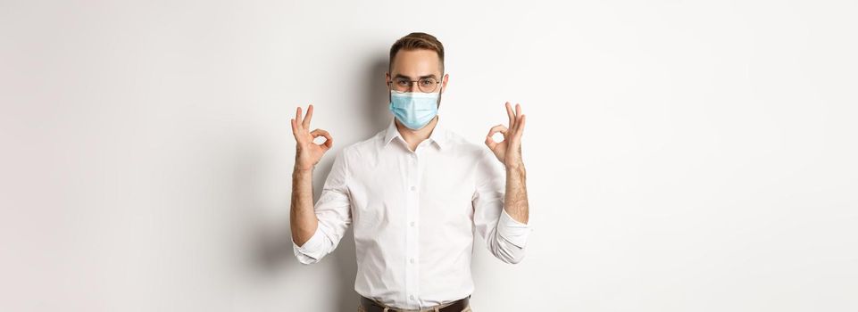 Covid-19, social distancing and quarantine concept. Confident businessman wearing medical mask and showing okay signs in approval, white background.