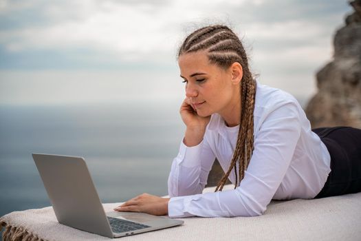 A woman is lying and typing on a laptop keyboard on a terrace with a beautiful sea view. Wearing a white blouse and black skirt. Freelance travel and vacation concept, digital nomad