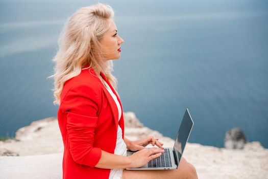 A woman is typing on a laptop keyboard on a terrace with a beautiful sea view. Freelancing, digital nomad, travel and vacation concept