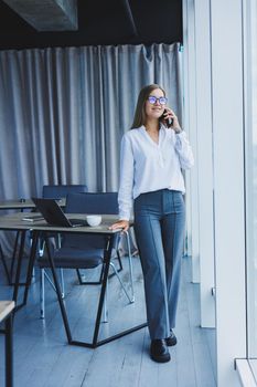 Photo of a successful young woman in a shirt smiling and working on a laptop while talking on the phone in a modern office with large windows. Remote work with coffee