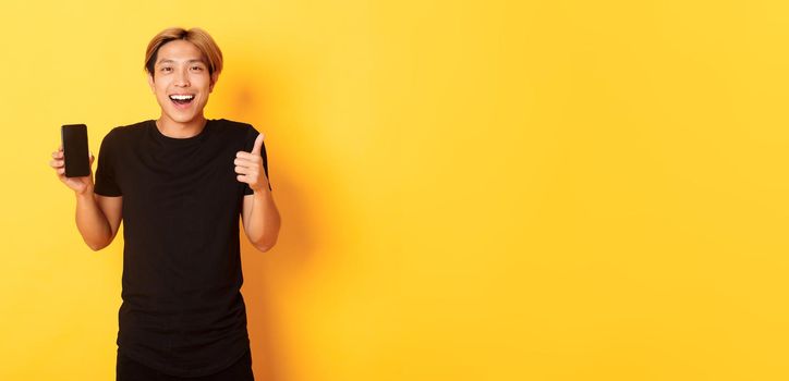 Portrait of pleased smiling asian guy, showing smartphone screen and thumbs-up satisfied with application, standing yellow background.