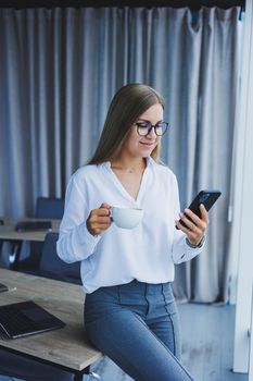 Photo of a successful young woman in a shirt smiling and working on a laptop while talking on the phone in a modern office with large windows. Remote work with coffee