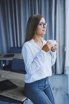Young smiling business woman standing in office near window with coffee in hands. A female manager in glasses and a white shirt is resting from work