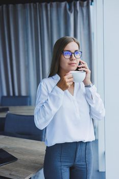 Photo of a successful young woman in a shirt smiling and working on a laptop while talking on the phone in a modern office with large windows. Remote work with coffee