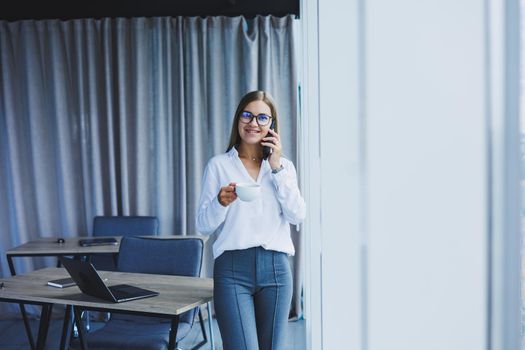Photo of a successful young woman in a shirt smiling and working on a laptop while talking on the phone in a modern office with large windows. Remote work with coffee