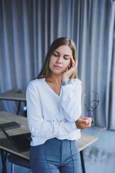 Business woman sitting at the desk in the office with a headache. A female manager in glasses and a white shirt suffers from a migraine