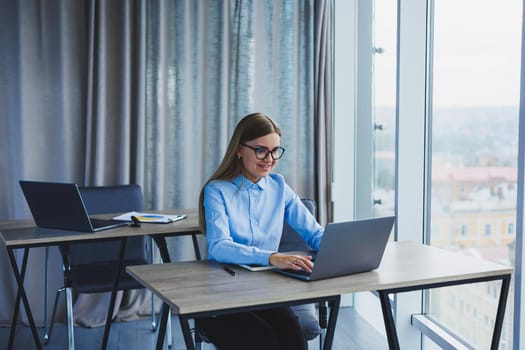 A beautiful woman in glasses sits at a table in the office and works at a laptop. Corporate work in the office. Modern woman workplace manager in classic clothes