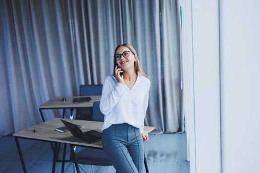 A successful smiling business woman is standing in the office by the window with a phone in her hands. A female manager in glasses and a white shirt is talking on the phone