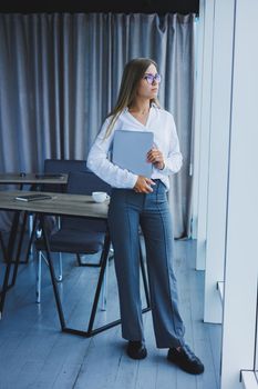 A young smiling business woman is standing in the office by the window with a laptop in her hands. Woman manager in glasses and white shirt