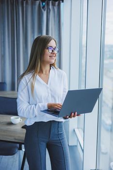 A young smiling business woman is standing in the office by the window with a laptop in her hands. Woman manager in glasses and white shirt