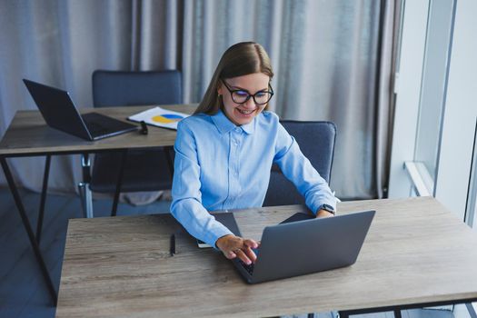 The secretary checks the schedule. Cheerful young beautiful woman in glasses with a smile uses a laptop while sitting at her workplace