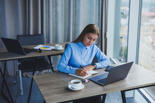 The secretary checks the schedule. Cheerful young beautiful woman in glasses with a smile uses a laptop while sitting at her workplace