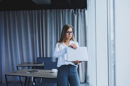 A young smiling business woman is standing in the office by the window with a laptop in her hands. Woman manager in glasses and white shirt