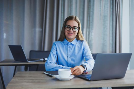 A beautiful business woman in a shirt and glasses sits with a laptop in the office at the table. Woman manager in glasses at workplace in modern office. Working day at the computer