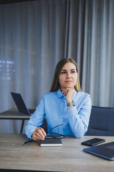 A beautiful woman in glasses sits at a table in the office and works at a laptop. Corporate work in the office. Modern woman workplace manager in classic clothes