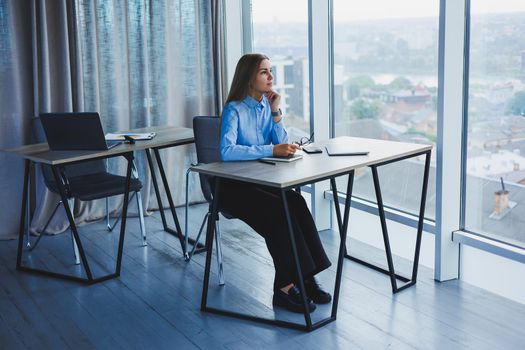 A beautiful woman in glasses sits at a table in the office and works at a laptop. Corporate work in the office. Modern woman workplace manager in classic clothes