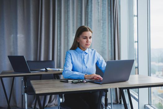 The secretary checks the schedule. Cheerful young beautiful woman in glasses with a smile uses a laptop while sitting at her workplace