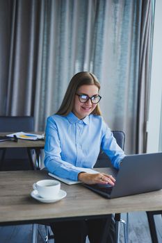 A beautiful business woman in a shirt and glasses sits with a laptop in the office at the table. Woman manager in glasses at workplace in modern office. Working day at the computer