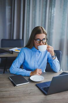 A beautiful business woman in a shirt and glasses sits with a laptop in the office at the table. Woman manager in glasses at workplace in modern office. Working day at the computer
