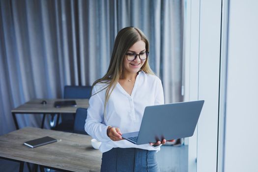 A young smiling business woman is standing in the office by the window with a laptop in her hands. Woman manager in glasses and white shirt