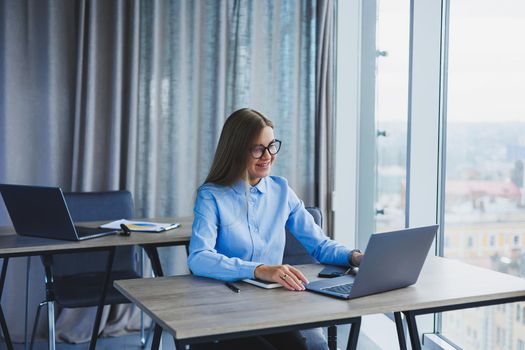 A beautiful woman in glasses sits at a table in the office and works at a laptop. Corporate work in the office. Modern woman workplace manager in classic clothes