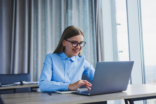 A beautiful woman in glasses sits at a table in the office and works at a laptop. Corporate work in the office. Modern woman workplace manager in classic clothes