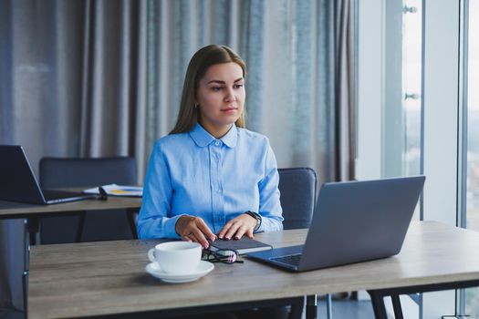 The secretary checks the schedule. Cheerful young beautiful woman in glasses with a smile uses a laptop while sitting at her workplace