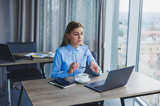 Adult smiling female manager in glasses and formal shirt sits at a desk and looks at a laptop. Modern, spacious, bright office with a large window.